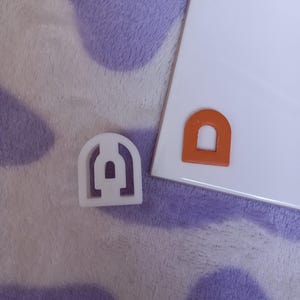 May include: Two cookie cutters in the shape of an archway. The cutter on the left is white plastic and the cutter on the right is orange plastic.