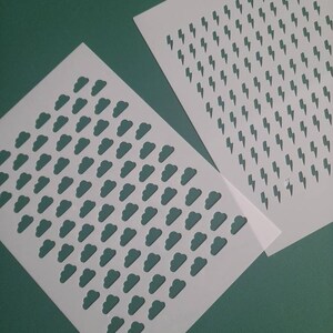 May include: Two white stencils, one with a repeating pattern of clouds and the other with a repeating pattern of lightning bolts.