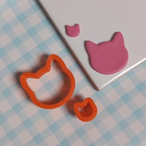 May include: Two orange plastic cat-shaped cookie cutters on a blue and white checked surface. The larger cutter is in the foreground and the smaller cutter is in the background.