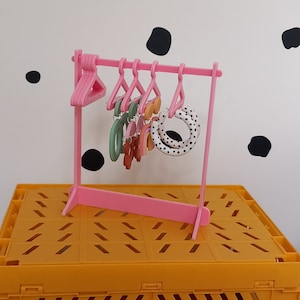 May include: A pink miniature clothes rack with a set of colourful earrings hanging from it. The rack is on a yellow surface.