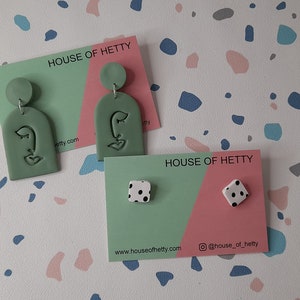 May include: Two pairs of earrings on cards with the text "HOUSE OF HETTY". The first pair is green with a face design. The second pair is white with black polka dots.