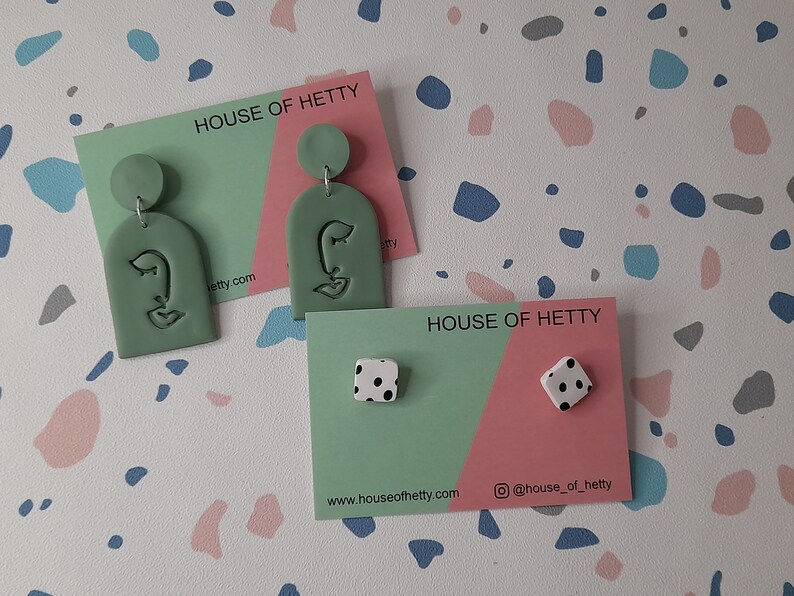 May include: Two pairs of earrings on cards with the text "HOUSE OF HETTY". The first pair is green with a face design. The second pair is white with black polka dots.