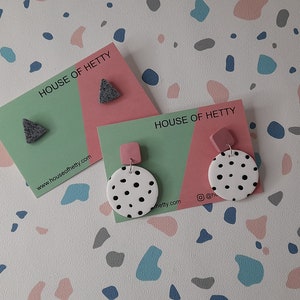 May include: Two pairs of earrings on cards with the text "HOUSE OF HETTY" and the website address "www.houseofhetty.com". The first pair is a gray triangle shape. The second pair is a white circle with black polka dots and a pink square.