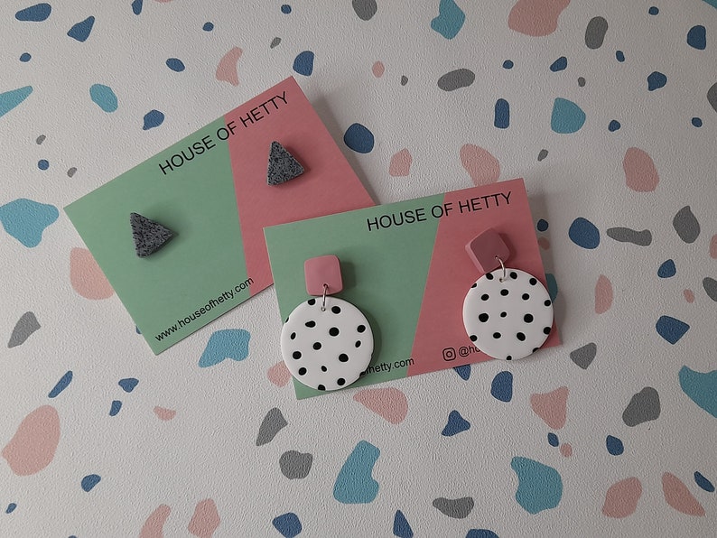 May include: Two pairs of earrings on cards with the text "HOUSE OF HETTY" and the website address "www.houseofhetty.com". The first pair is a gray triangle shape. The second pair is a white circle with black polka dots and a pink square.