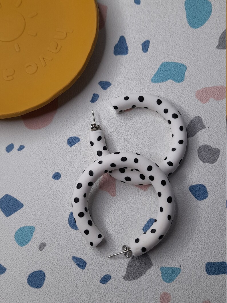 May include: A pair of white hoop earrings with black polka dots. The earrings are made of a thick, rounded material.