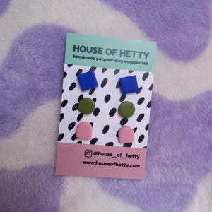May include: A card displaying six pairs of handmade polymer clay earrings in blue square, olive green circle, and pink half-circle shapes. The card has a black and white polka dot pattern and the text "HOUSE OF HETTY" and social media information.