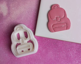 May include: A white plastic cookie cutter shaped like a pink backpack. The cookie cutter is on a pink sparkly surface.