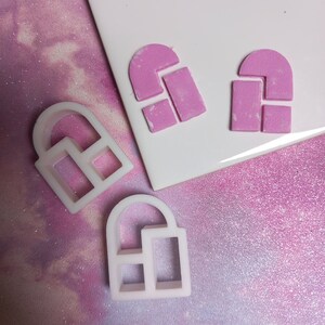 May include: Two pink plastic molds shaped like arches with a square cutout in the middle, and two white plastic molds shaped like arches with a square cutout in the middle, on a pink and white glittery surface.