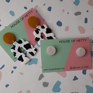May include: Two sets of earrings on cards with the text "House of Hetty". The first set features a white and black cow print design with a mustard yellow circle. The second set features two white round earrings.
