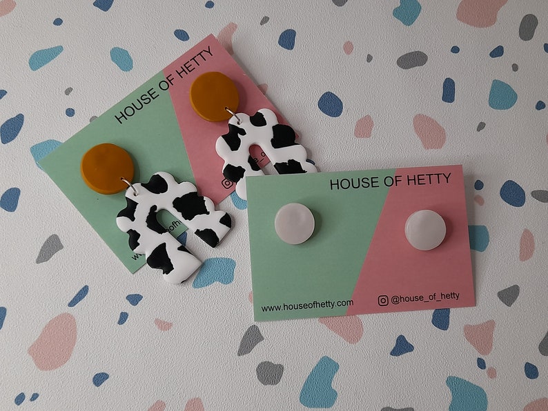 May include: Two sets of earrings on cards with the text "House of Hetty". The first set features a white and black cow print design with a mustard yellow circle. The second set features two white round earrings.