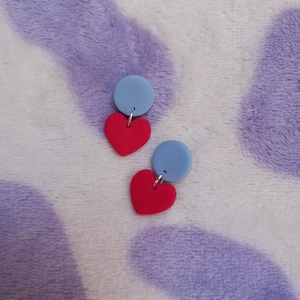 May include: A pair of earrings featuring a red heart shape suspended from a light blue circle. The earrings are made of a smooth material and hang from silver-colored hooks. The earrings are displayed on a purple and white patterned surface.