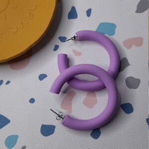 May include: A pair of large, purple hoop earrings made of a thick, matte material. The earrings are simple and modern in design.