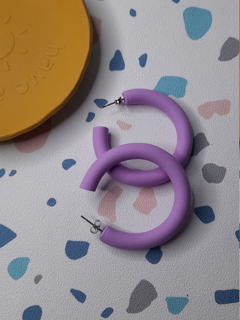 May include: A pair of large, purple hoop earrings made of a thick, matte material. The earrings are simple and modern in design.