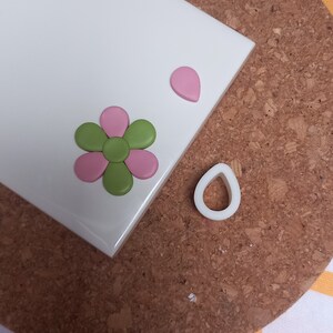 May include: A white tile with a pink and green flower made of polymer clay. There is a pink teardrop shape and a white teardrop-shaped cutter on the tile.