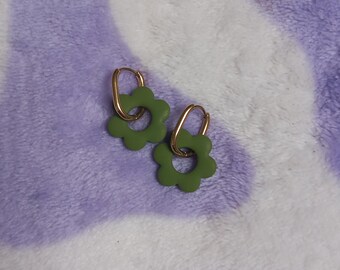 May include: A pair of gold hoop earrings with olive green flower-shaped charms. The earrings are displayed on a soft, purple and white patterned surface. The flower charms have a simple, minimalist design.