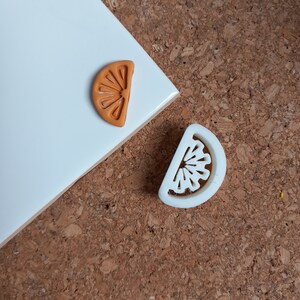 May include: Two orange slice shaped cookie cutters, one white and one orange. The cutters are on a cork surface.