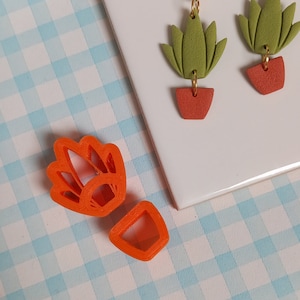 May include: Two orange plastic cookie cutters, one shaped like a plant with leaves and the other shaped like a pot.