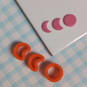 May include: Three orange plastic cookie cutters shaped like a crescent moon and a circle. Three pink fondant shapes are on a white surface.