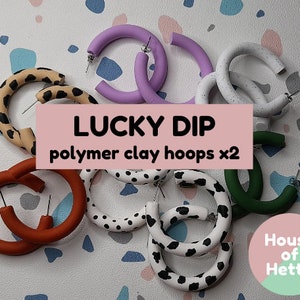 May include: A set of eight colorful polymer clay hoop earrings in various patterns, including leopard print, polka dots, and cow print. The earrings are arranged on a white and gray tiled surface. The text "LUCKY DIP polymer clay hoops x2" is displayed on a pink banner. The text "House of Hetty" is displayed on a pink and green circle.