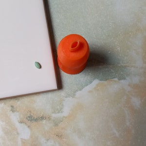 May include: An orange, cylindrical object with a small, oval opening on top. A small, green, oval-shaped object is next to a white rectangular object. The objects are on a surface with a marbled pattern.