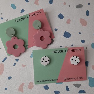 May include: Two sets of earrings on display cards. The first set features pink flower-shaped earrings with a green circle on top. The second set features white hexagonal earrings with black polka dots. The cards are green and pink and feature the text "House of Hetty" and the website address "www.houseofhetty.com".