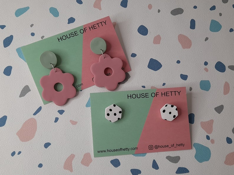 May include: Two sets of earrings on display cards. The first set features pink flower-shaped earrings with a green circle on top. The second set features white hexagonal earrings with black polka dots. The cards are green and pink and feature the text "House of Hetty" and the website address "www.houseofhetty.com".