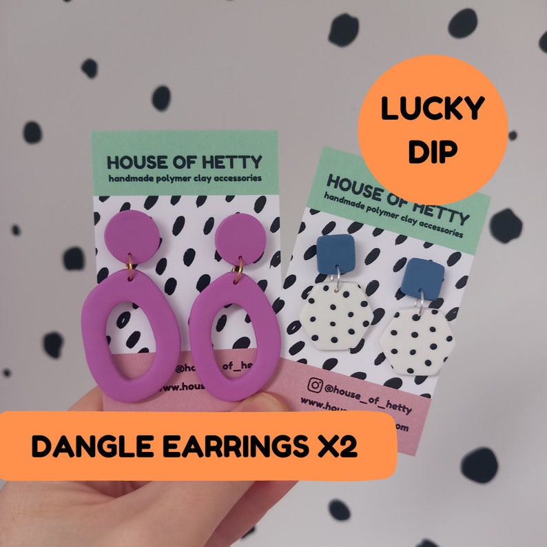May include: Two pairs of dangle earrings. The first pair is purple with a rounded oval shape. The second pair is white with black polka dots and a hexagonal shape. Both pairs are handmade polymer clay earrings. The cards say "House of Hetty" and "Lucky Dip".
