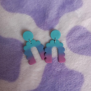 May include: A pair of ombre arch-shaped earrings with a pink, blue, and turquoise color gradient. The earrings have a scalloped edge and are made of polymer clay.