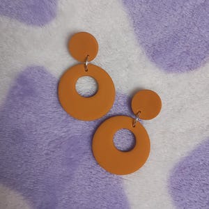 May include: A pair of orange earrings. Each earring has a small, round stud connected to a larger, circular pendant with a hole in the centre. The earrings are a warm, earthy orange colour.