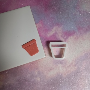 May include: A white plastic cookie cutter shaped like a flower pot, next to a small, terracotta-coloured flower pot.