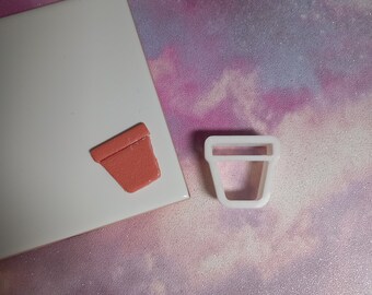 May include: A white plastic cookie cutter shaped like a flower pot, next to a small, terracotta-colored flower pot.