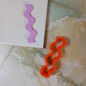 May include: Two wavy cookie cutters, one pink and one orange, on a white tile surface.