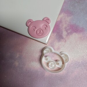 May include: A pink clay bear face and a white plastic bear cookie cutter. Both are on a sparkly purple and white background.
