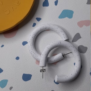 May include: A pair of white hoop earrings with a speckled texture. The earrings are made of a thick, rounded material and have a silver post.