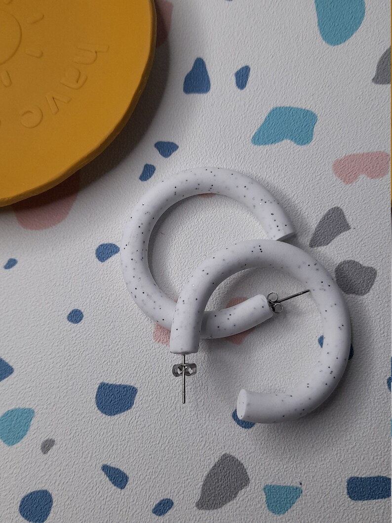 May include: A pair of white hoop earrings with a speckled texture. The earrings are made of a thick, rounded material and have a silver post.