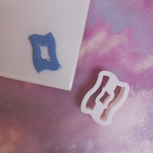 May include: Two cookie cutters, one blue and one white, shaped like a wavy rectangle with a smaller rectangle cut out in the centre.