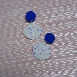 May include: A pair of earrings with a white seashell design, speckled with blue, and a solid blue circle above. The earrings are presented on a light pink, textured surface.