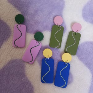 May include: A collection of colorful, abstract earrings. The earrings feature a circular top and a rectangular bottom with a wavy line design. Colors include pink, green, blue, and yellow. The earrings are displayed on a purple and white patterned surface.