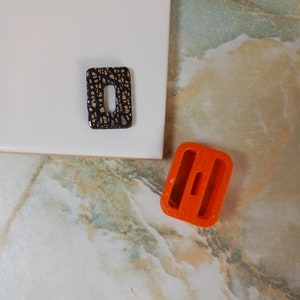 May include: Two differently shaped, decorative, ceramic tiles. One tile is black with gold flecks and the other is orange.