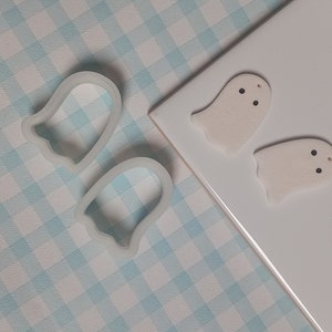 May include: Two white ghost-shaped ornaments with black eyes against a white background. Two clear plastic ghost-shaped cookie cutters are also shown.