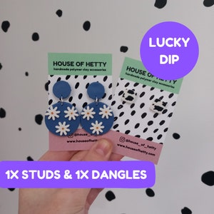 May include: Two pairs of handmade polymer clay earrings. One pair features blue and white daisy flower dangles. The other pair features black and white cow print studs. The cards have the text "House of Hetty" and "Lucky Dip".