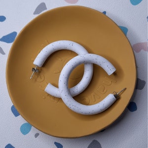 May include: A pair of white hoop earrings with a speckled texture. The earrings are on a yellow plate.