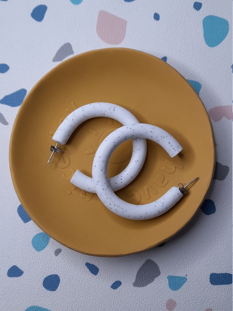 May include: A pair of white hoop earrings with a speckled texture. The earrings are on a yellow plate.