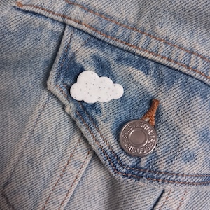May include: A white cloud-shaped pin with black speckles on a blue denim jacket with a silver button that says "Levi's" on it.