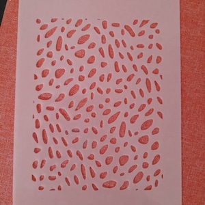 May include: A pink plastic stencil with a repeating pattern of small, irregular shapes cut out. The shapes are reminiscent of pebbles or small stones.