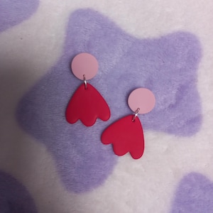 May include: A pair of handmade earrings. Each earring has a light pink disc stud and a vibrant red, petal-shaped drop. The earrings hang from silver-coloured hooks, set against a soft, patterned background.