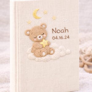 May include: Cream-colored baby memory book with an embroidered teddy bear holding a star on the cover. The cover also features a crescent moon, stars, and the name "Noah" with the date "04.16.24".