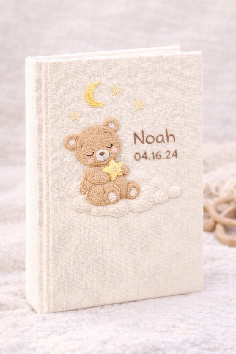 May include: Cream-colored baby memory book with an embroidered teddy bear holding a star on the cover. The cover also features a crescent moon, stars, and the name "Noah" with the date "04.16.24".