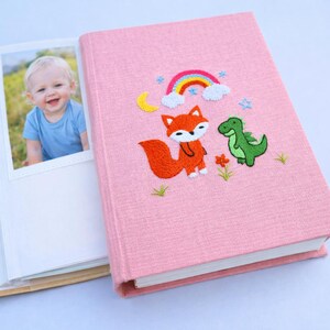 May include: Pink photo album with embroidered design featuring a fox, dinosaur, rainbow, moon, and flowers. The album is open, revealing a photo of a smiling baby. The album is made of a textured fabric.