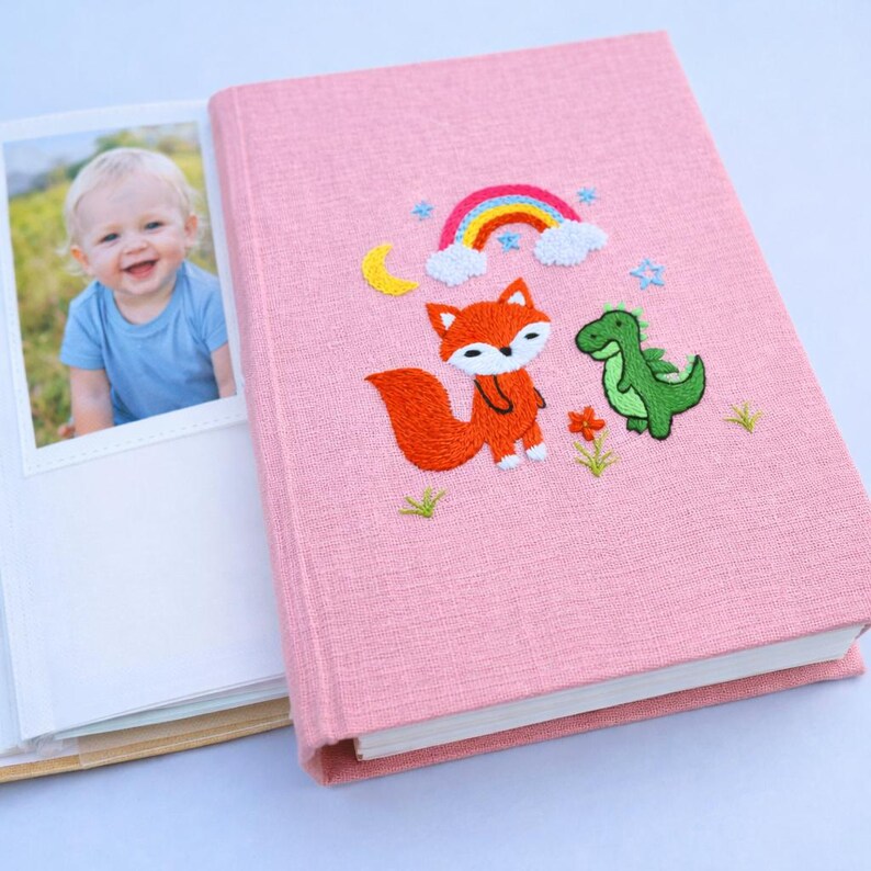 May include: Pink photo album with embroidered design featuring a fox, dinosaur, rainbow, moon, and flowers. The album is open, revealing a photo of a smiling baby. The album is made of a textured fabric.